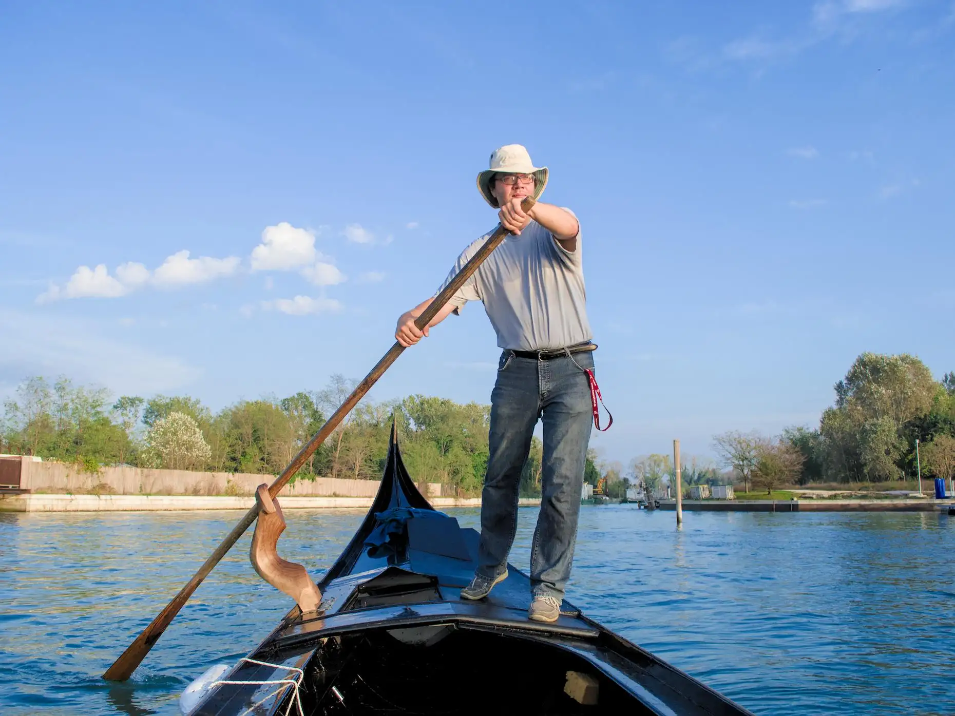 A man in jeans, t-shirt and a hat, on the back of a Venetian gondola, rowing. In the background, several islands with trees.